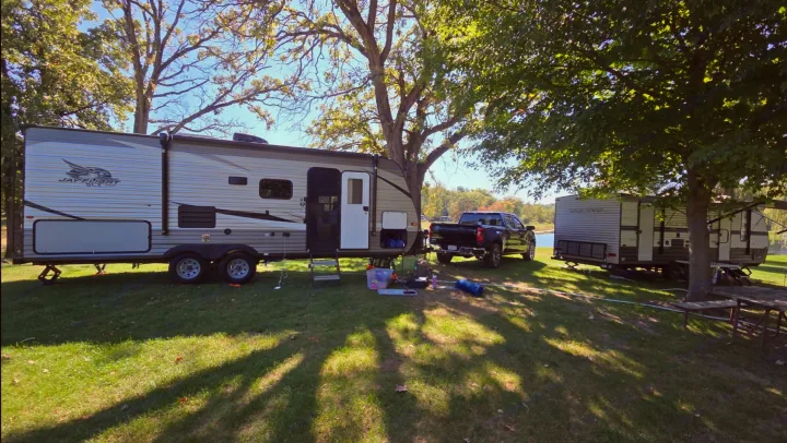 Two travel trailer RVs and pickup trucks are parked on grass by a lake, shaded by large trees for a relaxing RV rental camping trip.. Jayco Jay Flight SLX 2021