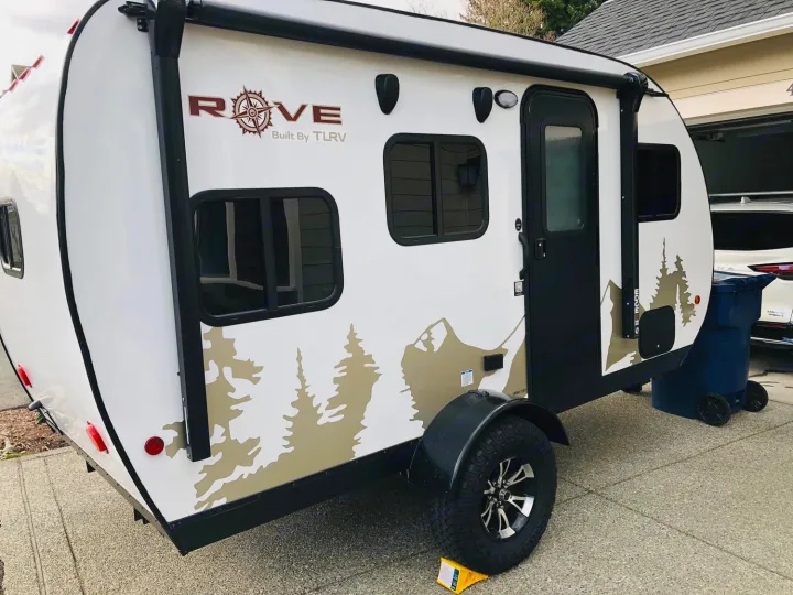 This white Rove compact RV rental is parked on a driveway. It features nature decals, an awning, black door & windows.. Travel Lite Rove Lite 2021