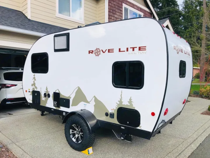 White Rove Lite travel trailer, ideal for rv rental. Features outdoor graphics, rugged wheels, & utility access, parked on a driveway.. Travel Lite Rove Lite 2021
