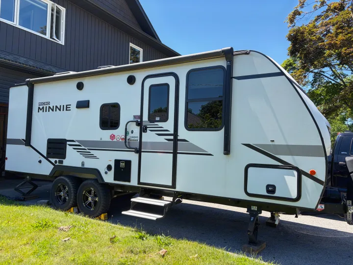 White Winnebago Micro Minnie travel trailer, ideal for an rv rental, parked next to a dark house on a sunny day.. Minnie Micro Minnie 2021