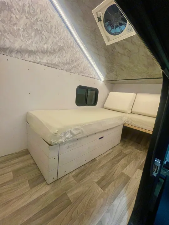 Cozy RV rental interior featuring a white mattress bed with storage, L-shaped seating, small window, and wood-look floor.. Homemade Box 2020