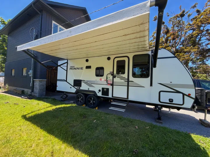 White Micro Minnie travel trailer RV rental with extended awning, parked on gravel next to a dark house.. Minnie Micro Minnie 2021