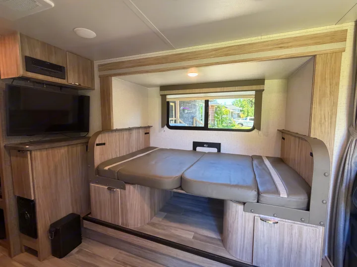 RV rental interior featuring a grey cushioned bed/couch, entertainment center with TV, and a window revealing an outdoor scene.. Minnie Micro Minnie 2021