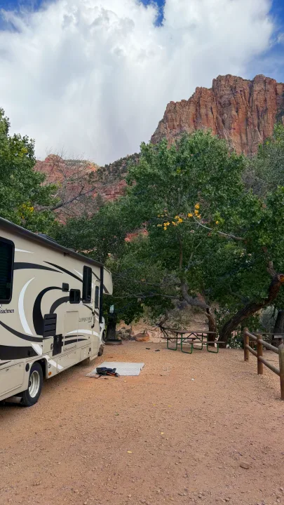 Scenic RV rental parked at a serene campsite with a picnic table, surrounded by lush trees and towering red mountains.. Coachmen Leprechaun 2021