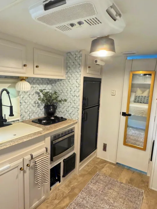 Modern RV rental kitchen with light cabinets, mosaic backsplash, compact fridge, microwave, and a mirror reflecting the bedroom.. Coachmen Apex 2017