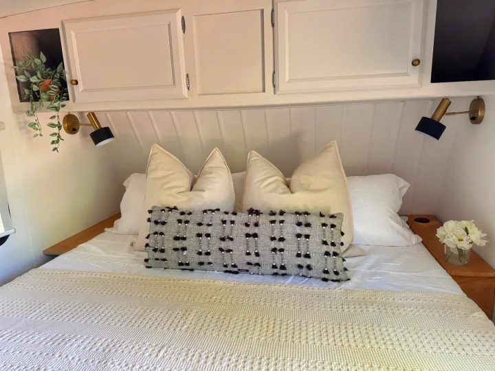 Cozy RV rental bedroom with white bedding, cream pillows, a textured grey pillow, brass wall lights, and plant details.. Coachmen Apex 2017