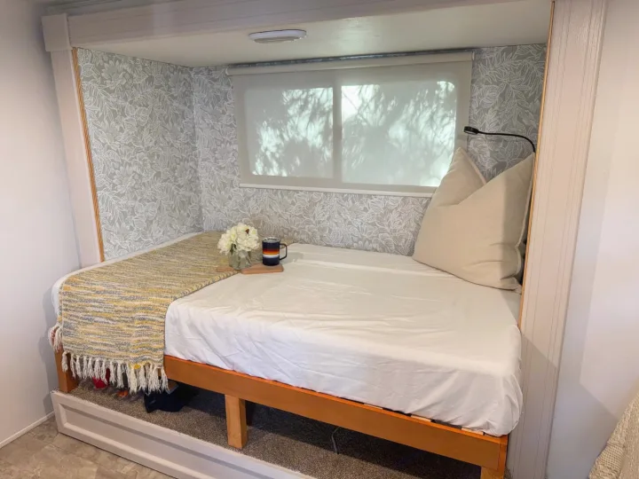 Comfortable RV rental bed nook with leaf-patterned wallpaper, soft bedding, and ambient window light.. Coachmen Apex 2017
