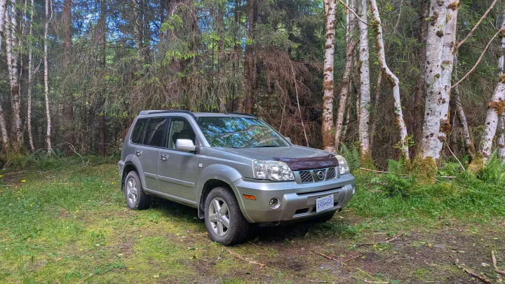 Silver SUV on a dirt path in a forest. This vehicle is great for outdoor trips; explore RV rental for more space.. Nissan Xtrail 2005