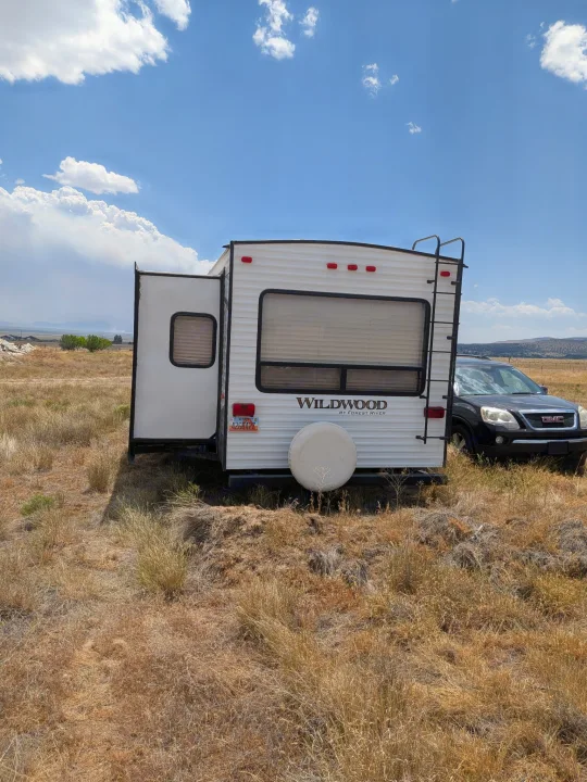 A white Wildwood travel trailer RV rental with an extended side is parked in a dry field, with a black GMC SUV nearby.. Forest River Wildwood 2017