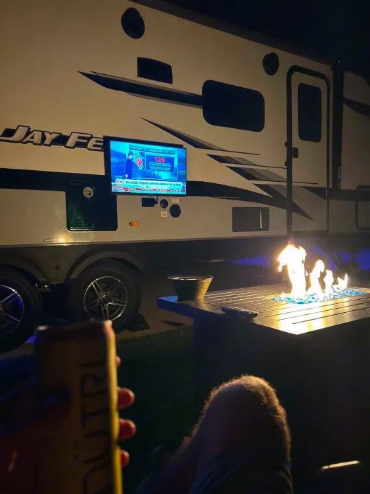 Relaxing outdoors in an RV rental. A TV shows storm news while blue flames flicker from a fire pit on a dark evening.. Jayco Jay Feather 2023