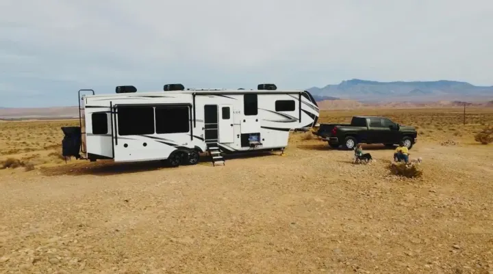 An RV rental fifth wheel and pickup truck parked in a vast desert landscape, with two people and their dogs relaxing outside.. Grand Design Solitude 2024