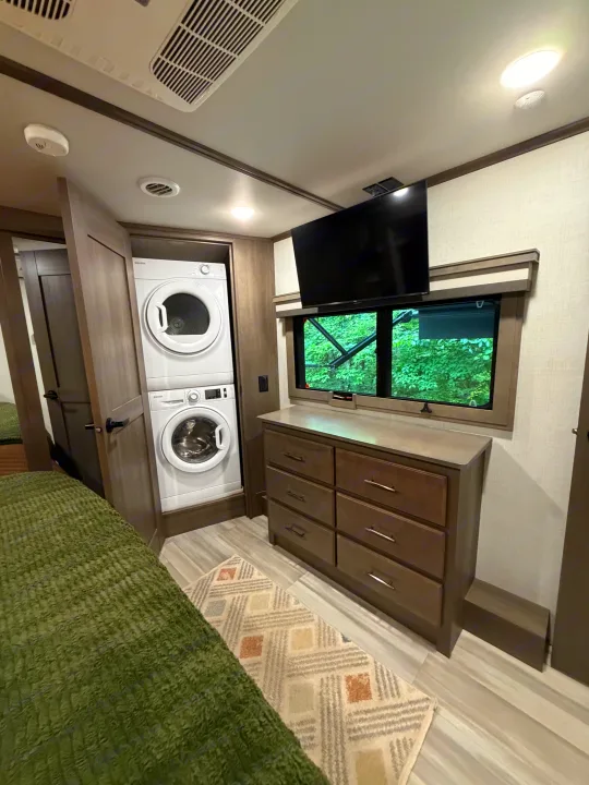 RV rental bedroom with stacked washer/dryer, dresser, TV, and a window overlooking green trees. Features a green bedspread and rug.. Grand Design Solitude 2024