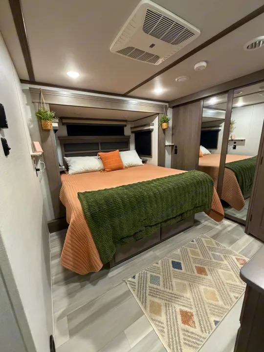 Modern RV rental bedroom: cozy bed with peach and green linens, mirrored closet, patterned rug, and ceiling AC.. Grand Design Solitude 2024