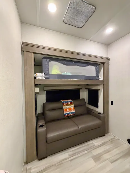 Inside a cozy RV rental: bunk beds with safety rail above a brown sofa with blanket and cup holders.. Grand Design Solitude 2024