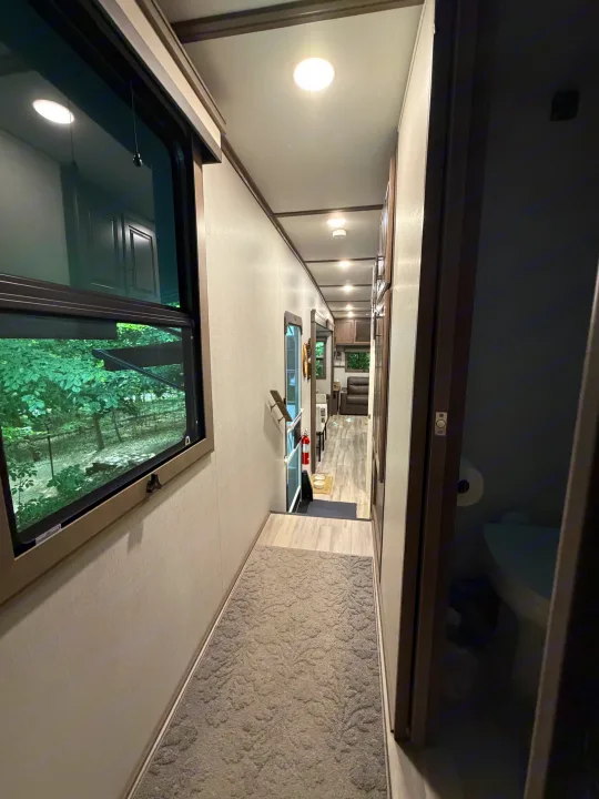 Well-lit RV rental hallway with recessed lights, a patterned rug, window to trees, and views into the living area and bathroom.. Grand Design Solitude 2024