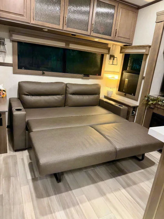 This RV rental features a versatile living area with a modern taupe pull-out sofa bed, extended for sleeping. Overhead cabinets and light wood flooring complete the interior.. Grand Design Solitude 2024