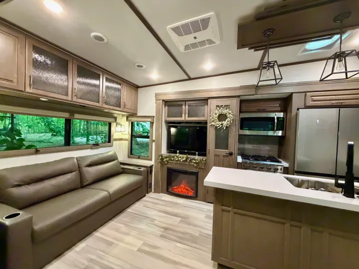 This modern RV rental interior features a comfortable living area with a sofa, a full kitchen, an electric fireplace, and scenic forest views.. Grand Design Solitude 2024