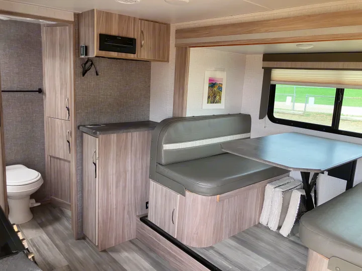 This rv rental features a modern interior with a dinette, kitchen, and toilet. Light wood tones, grey seating, and a window view.. Winnebago Micro Winnie 2021