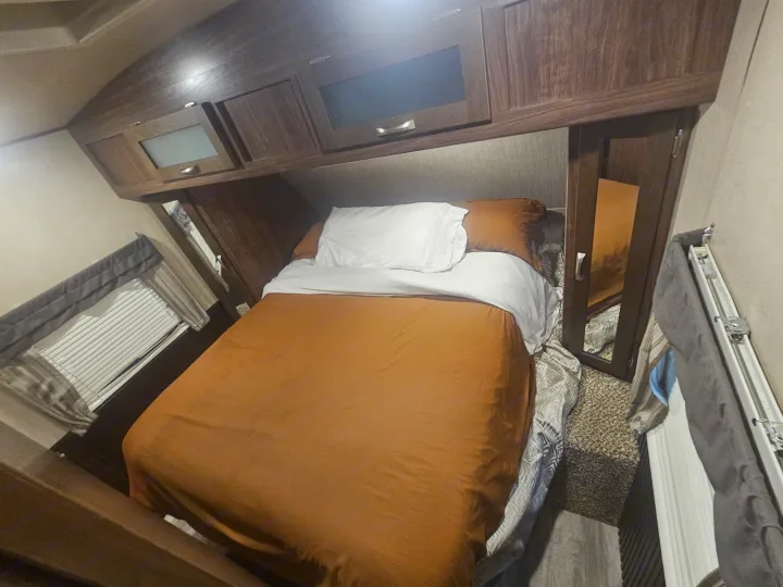 Cozy rv rental bedroom with an inviting orange comforter, white pillows, overhead cabinets, and a window with blinds.. Dutchmen 242bhsl 2017