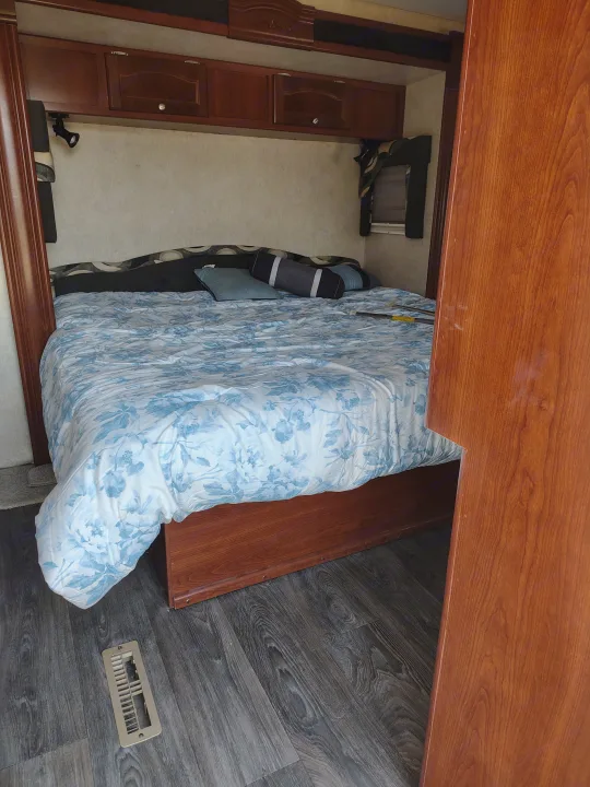 Compact RV rental bedroom featuring a bed with a blue floral duvet, overhead wood cabinets, and dark laminate flooring.. Fleetwood Terry 2007