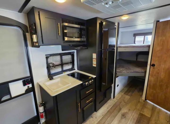 RV rental interior featuring a compact kitchen with sink, stove, microwave, fridge, and bunk beds, ideal for family travel.. Keystone RV Springdale 2023