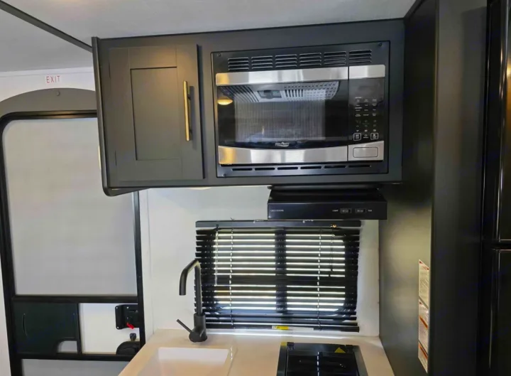 Modern RV rental kitchenette featuring dark cabinets, built-in microwave, sink, and stovetop with a window and blinds.. Keystone RV Springdale 2023