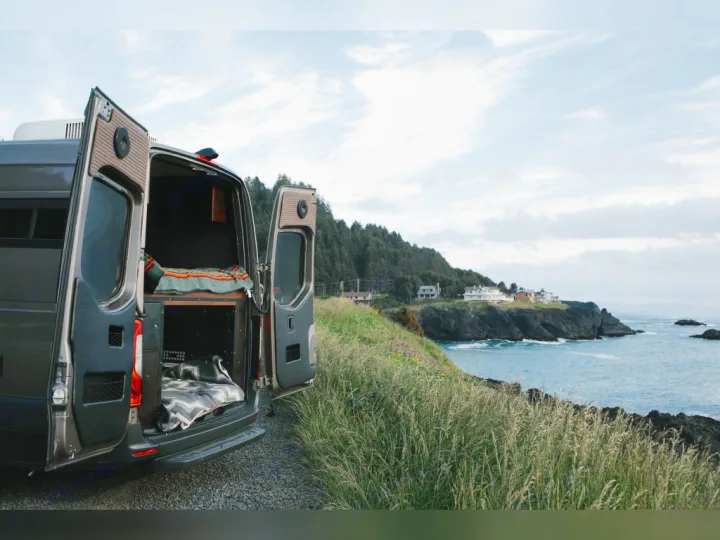 This RV rental, with its back doors open showing interior beds, is parked on a gravel path overlooking a scenic ocean vista with distant houses and cliffs.. Airstream Interstate 2025