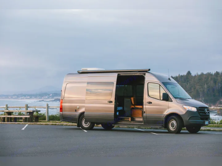 This RV rental camper van, with its door open, offers stunning ocean views from its parking spot near a forest.. Airstream Interstate 2025