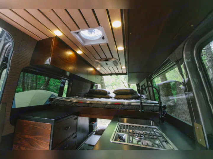 Van conversion RV rental interior: elevated bed, compact kitchen with stove/sink, and warm wooden ceiling. Ideal for nature trips.. Airstream Interstate 2025