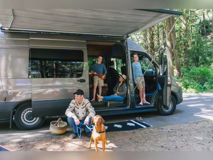 Family with dog by **RV rental** camper van in woods. Two boys play ukulele inside, a woman sits, and a man kneels with the dog.. Airstream Interstate 2025