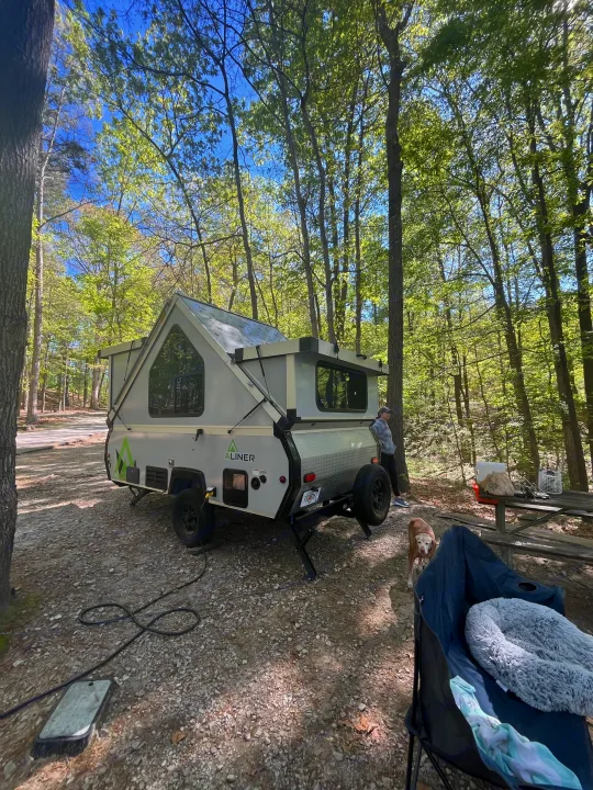 Enjoying a serene forest campsite with our RV rental, an A-liner pop-up camper, under a clear blue sky. A person and dog are nearby.. A-Liner Ranger 2025