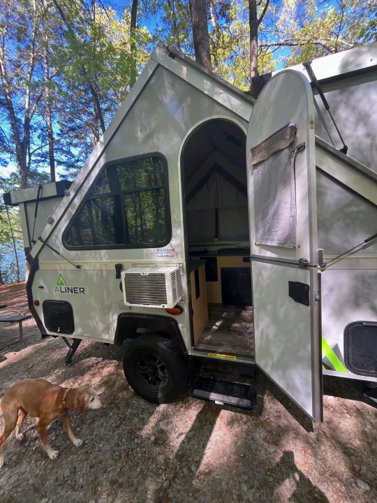 An A-Liner pop-up camper RV rental with an open door in a wooded area by water, with a dog.. A-Liner Ranger 2025