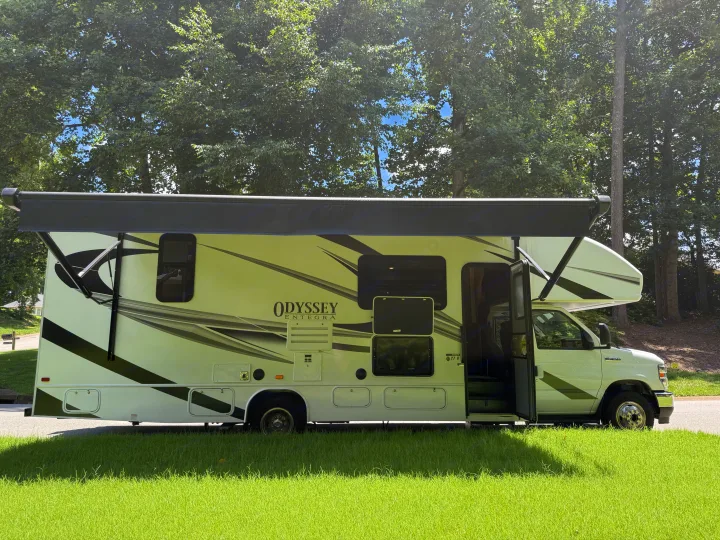 White Entegra Odyssey RV rental with awning extended, parked on asphalt with green grass. Trees overhead, door open.. Entegra Coach Odyssey 2023