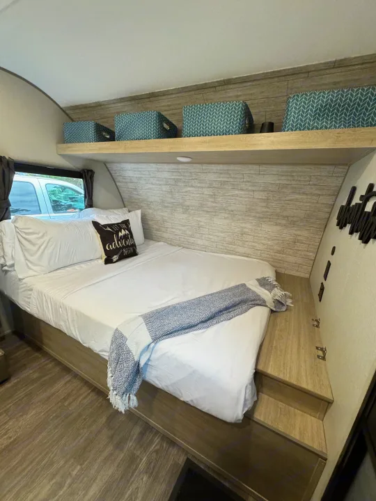 Cozy RV rental bedroom with white bedding, an "adventure" pillow, patterned throw, shelf storage, and wood accents.. Forest River Aurora Aurora Light 2026