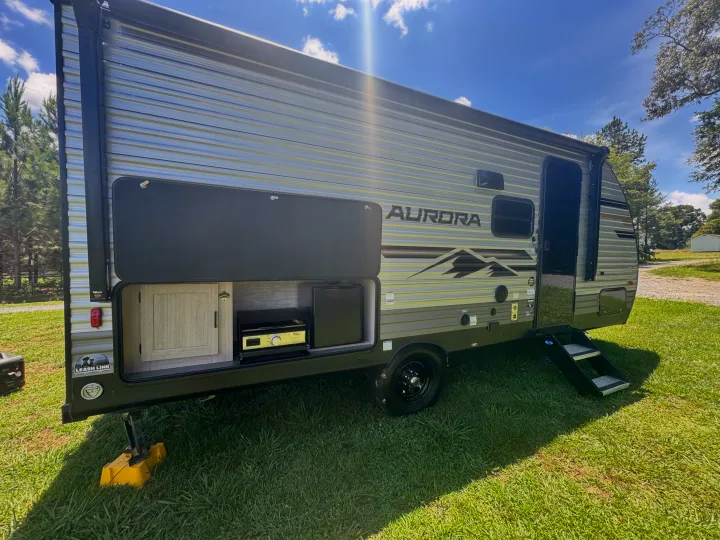This RV rental features an outdoor kitchen with a fridge and stove, perfect for camping adventures under a sunny sky.. Forest River Aurora Aurora Light 2026