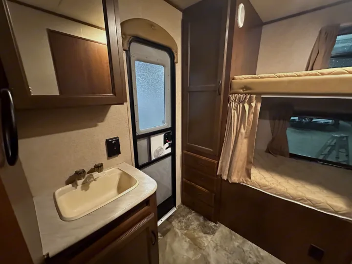 This RV rental interior showcases a compact sink, wooden cabinets, a main entrance door, and comfortable bunk beds with curtains.. Jayco Jay Flight 2015