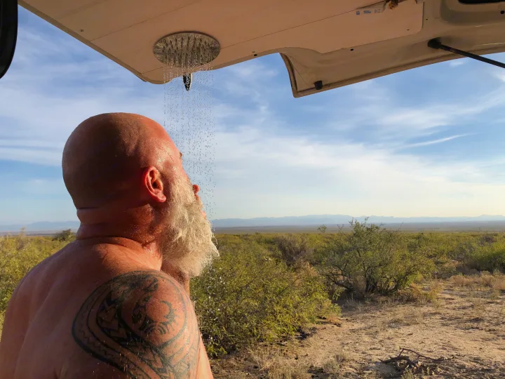 Outdoor shower from an RV rental: a bald man with a white beard and tattoo washes off in the desert landscape.. Winnebago Eurovan Camper Eurovan Camper 1997