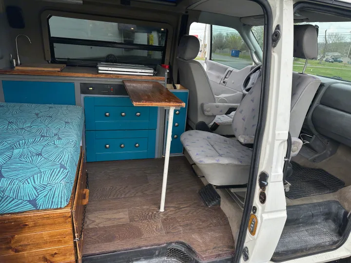 RV rental interior: custom camper van conversion with kitchenette, bed, storage, and a pull-out table. Front seats visible.. Winnebago Eurovan Camper Eurovan Camper 1997