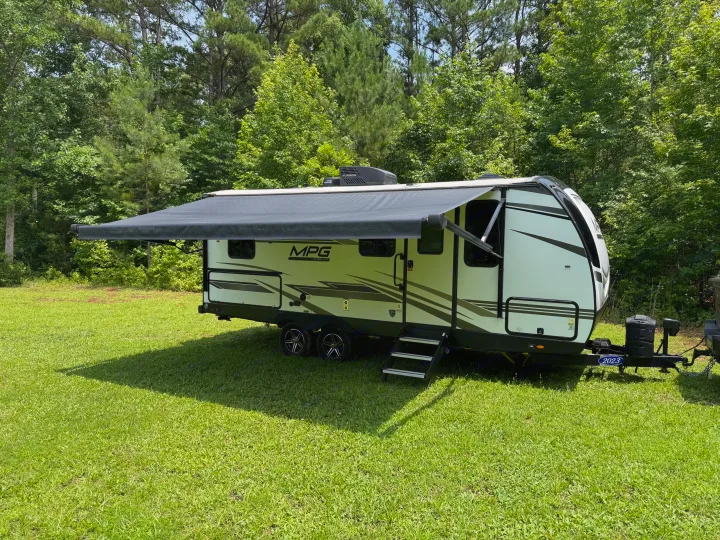 This RV rental features a modern travel trailer with its awning extended, parked on vibrant green grass bordered by lush trees.. Cruiser RV MPG 2023