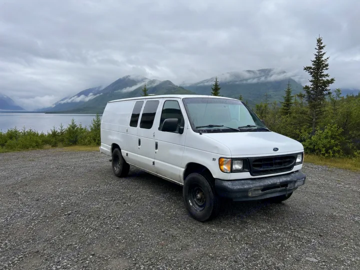 White Ford van, possibly an RV rental, parked on gravel overlooking a scenic lake and misty mountains. Perfect for van life adventures!. Ford Econolline 250s 1998