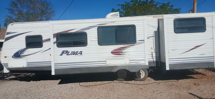 A large white Puma travel trailer for rv rental, featuring a slide-out, parked on gravel. Note the flat rear tire.. Palomino Puma 2013