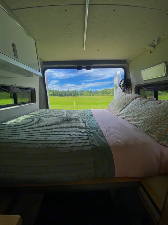 Relaxing RV rental bed with a view of a bright green field, forest, and blue sky from the open rear doors of a camper van.. Dodge Ram 1500 2017