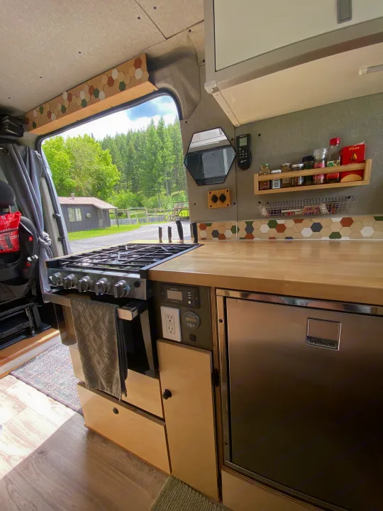 Spacious RV rental kitchen in a camper van with stove, fridge, wood counter, and a beautiful nature view outside.. Dodge Ram 1500 2017