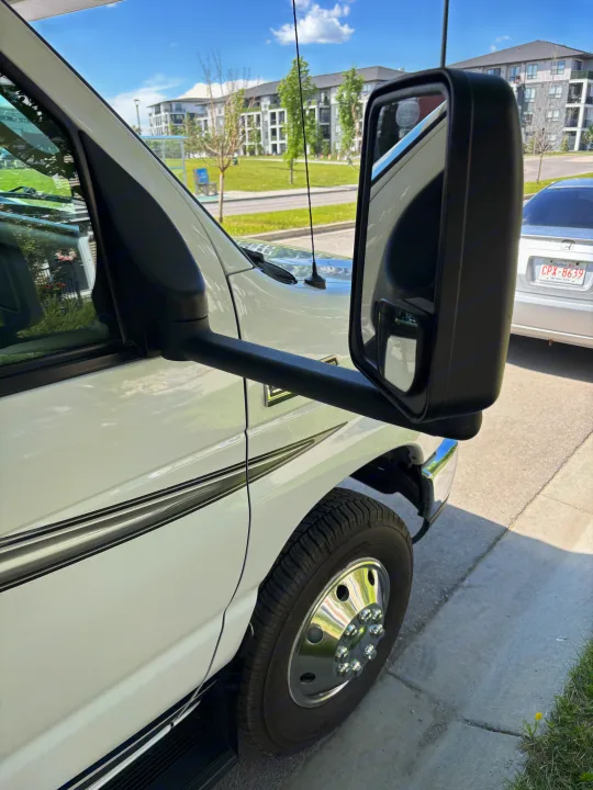 White RV rental van with large side mirror, reflecting a silver car. Apartments and street are visible in the sunny background.. Forest River Sunseeker LE 2020