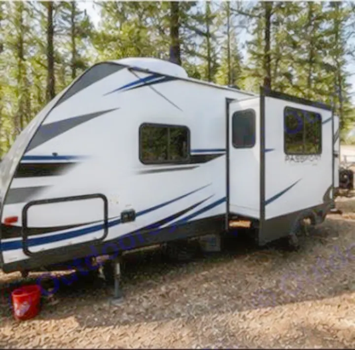 Explore the outdoors with this RV rental. A modern white travel trailer, door open, is parked in a wooded campsite.. Keystone RV Passport Ultra Lite 2019