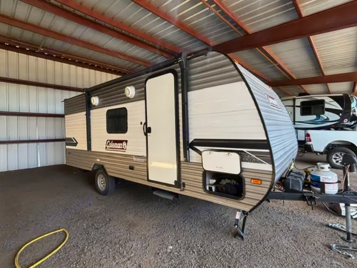 This Coleman travel trailer rv rental is neatly stored under a covered metal shed, featuring propane tanks and an open front compartment.. Dutchmen Coleman Lantern LT 2024
