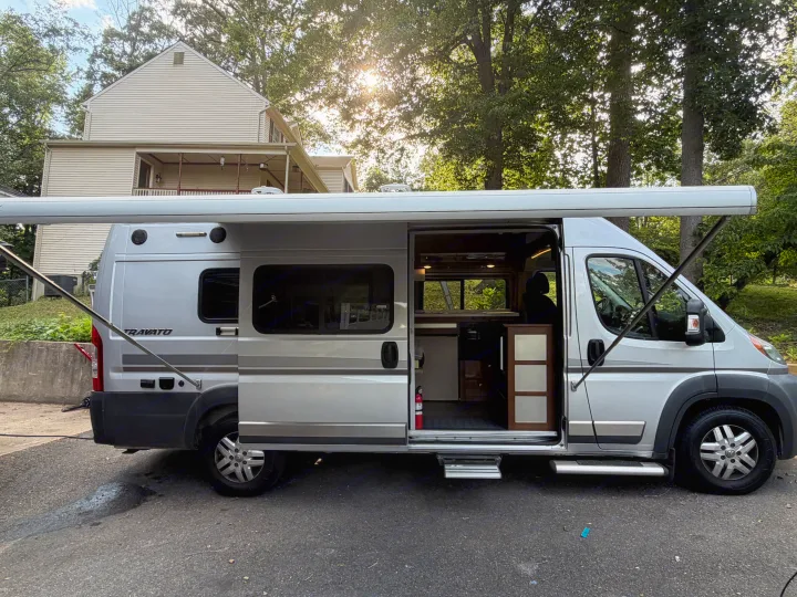 Silver Travato camper van, available for rv rental, with an extended awning and open side door revealing its furnished interior.. Winnebago Travato 2016