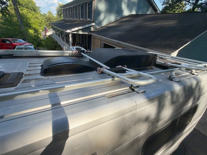 Silver RV rental rooftop: AC unit, roof rack, and vents visible, with a house, trees, and American flag in the background.. Winnebago Travato 2016