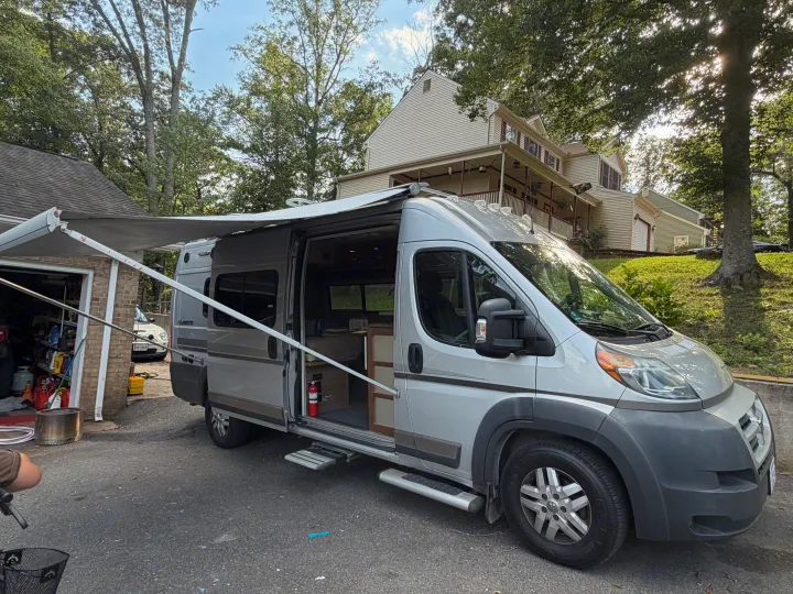 Silver RV rental with extended awning & open side door parked at a house, ready for adventure.. Winnebago Travato 2016