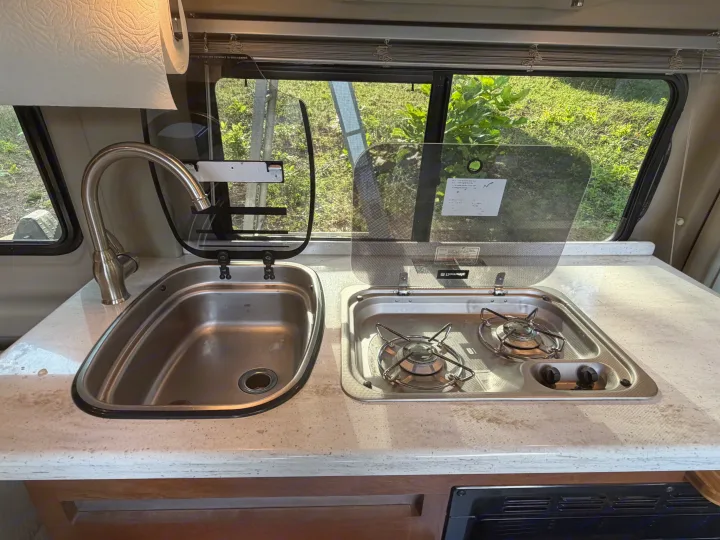This rv rental features a compact kitchen with a stainless steel sink, gooseneck faucet, and a two-burner stove set in a light countertop.. Winnebago Travato 2016